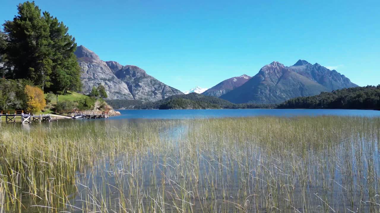 A low-flying drone glides over a lake, showcasing majestic mountains with a snow-capped peak under a radiant sun and blue sky