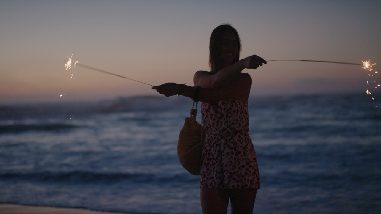 일몰 슬로우 모션 장난스러운 축하의 아름다운 해변에서 함께 새해 전야를 축하하며 춤추는 폭죽을 들고 있는 여자친구의 사진을 찍는 젊은 남자