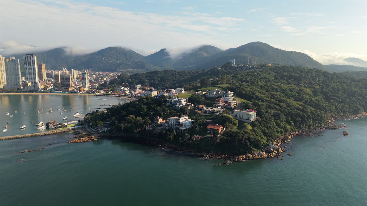 Backward aerial fly from Canto da Praia peninsula with lush green hills and Itapema's city skyline in background, Santa Catarina, Brazil
