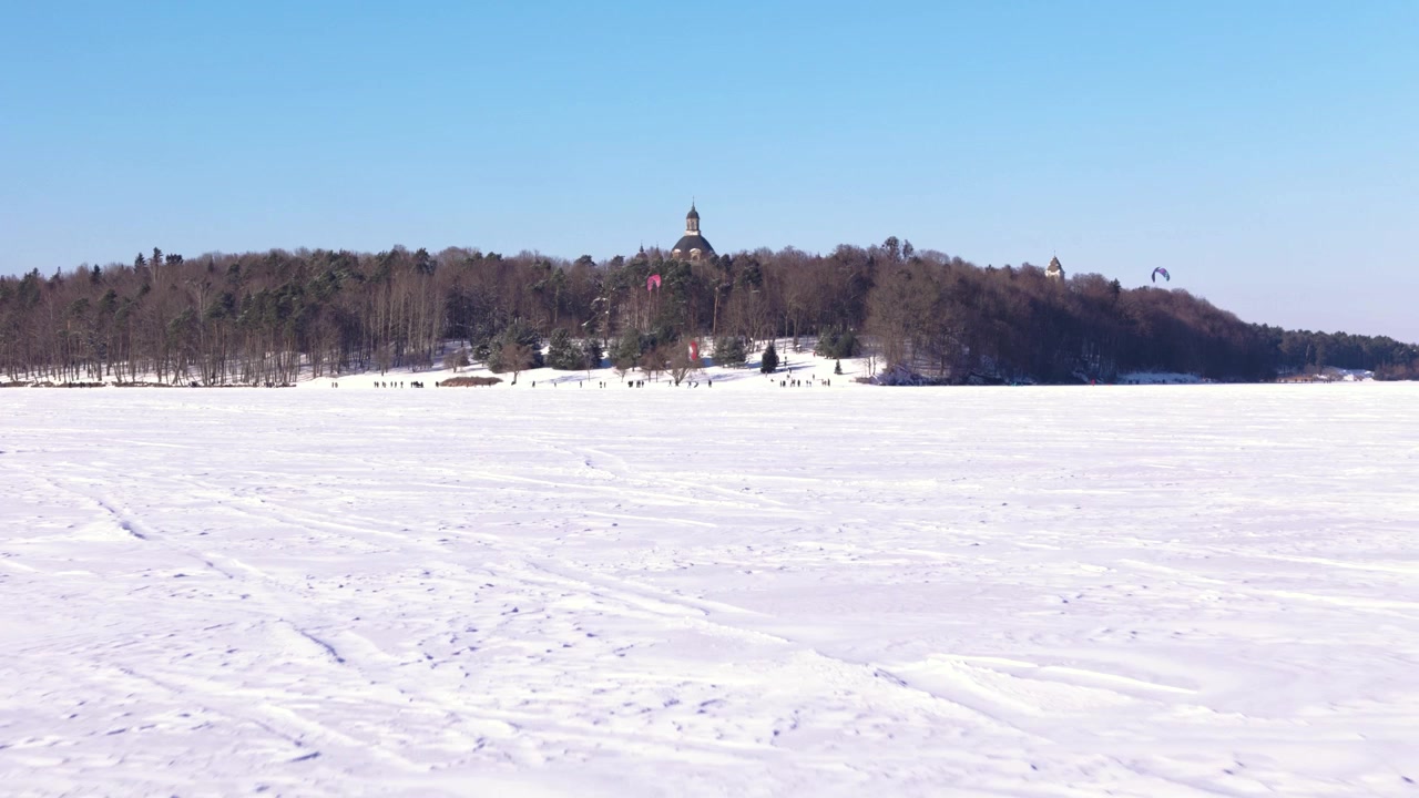 Sneklædt frossen sø med fjern bygning på skovklædt bred under klar blå himmel