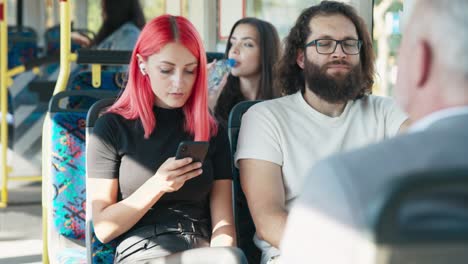 woman with pink hair rides public transportation bus, girl uses smartphone, has wireless in earphones