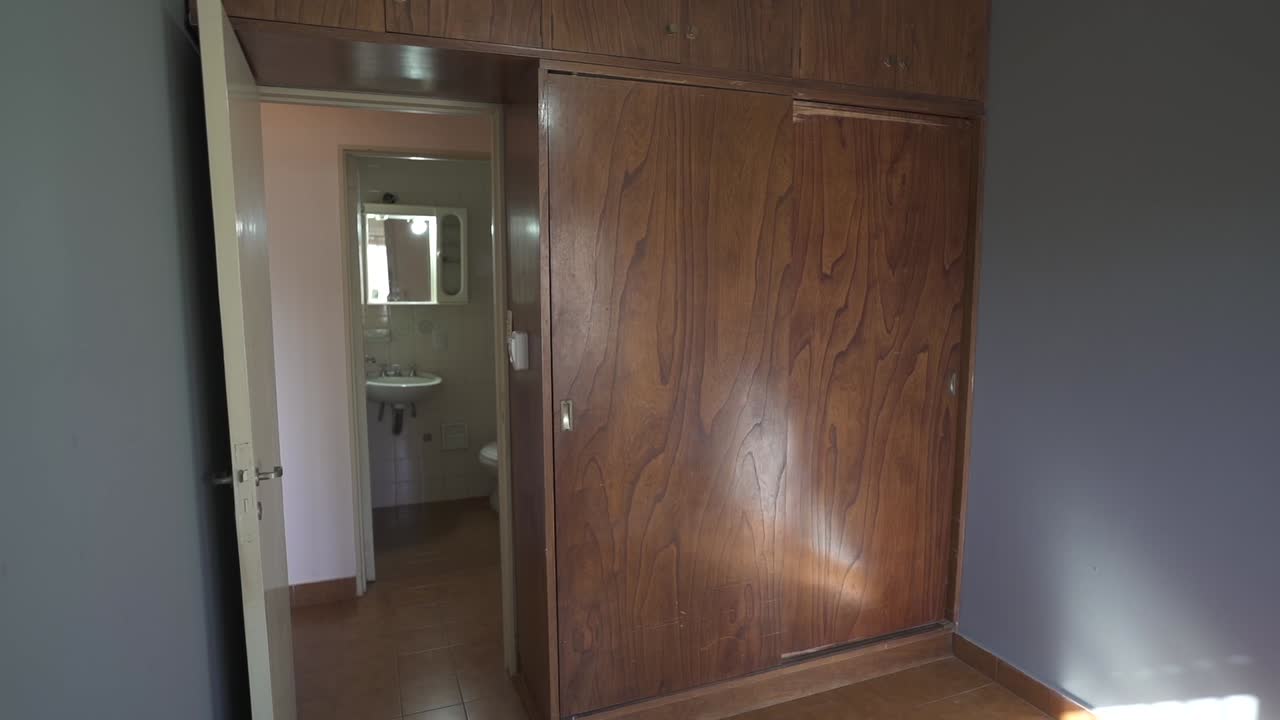 Slow, right motion camera clip portraying empty apartment room for real estate purposes, with large wooden closet and ceramic floor, and restroom in background.
