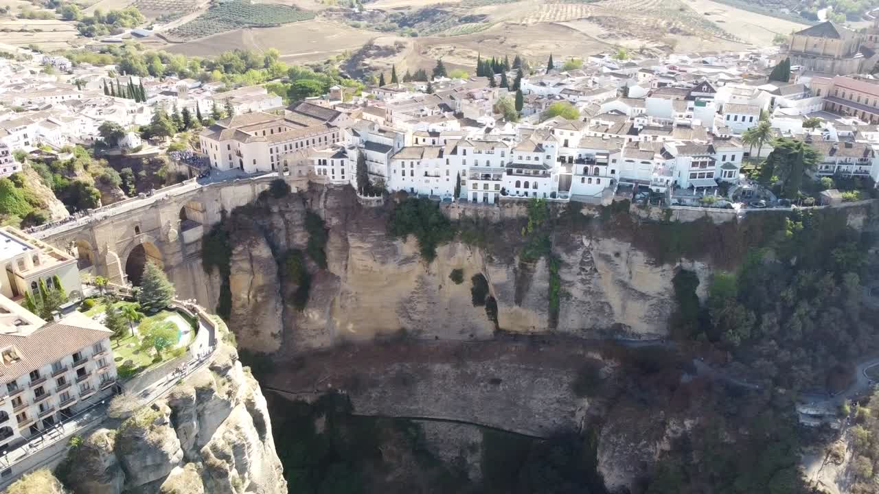 Aerial drone shot captures whitewashed buildings and historic bridge spanning deep gorge with dramatic cliffs in Ronda, Spain during sunny day offering stunning architecture and natural contras