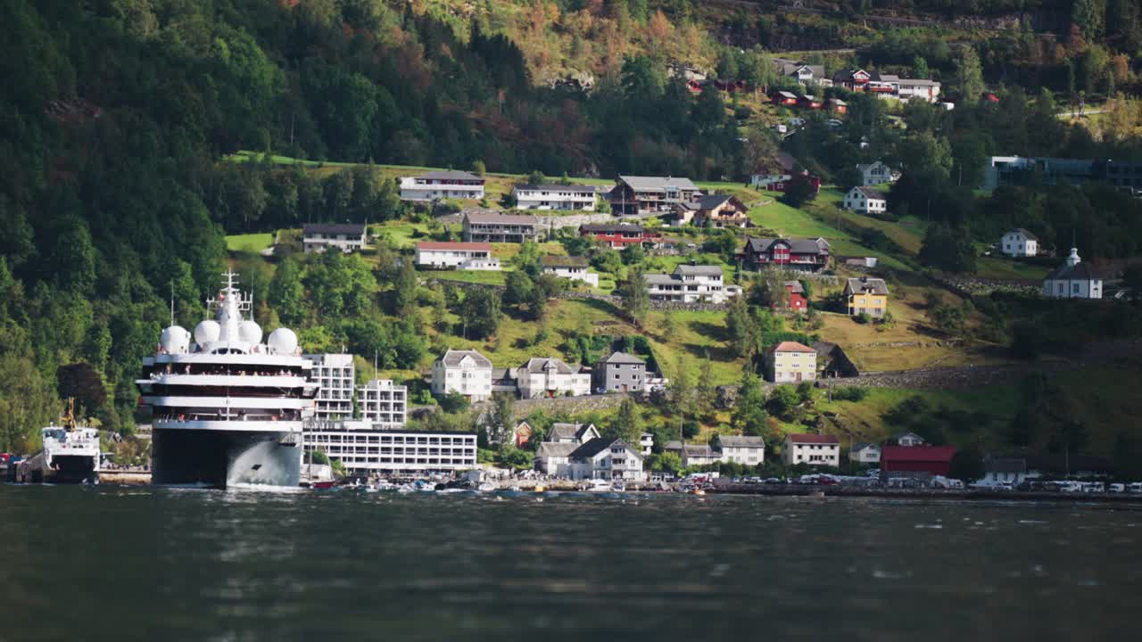 큰 크루즈 선박이 게이랑거 피오르드 마을 근처의 항구에서 출발합니다. 배경에는 집과 초록색 언덕이 있습니다.