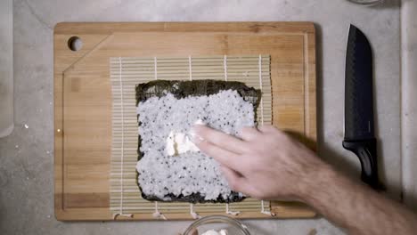 top down view of two hands on wooden cutting tablet on kitchen counter spreading cream with fingers on the chai seed rice on a mat and seaweed nori sheet to make sushi