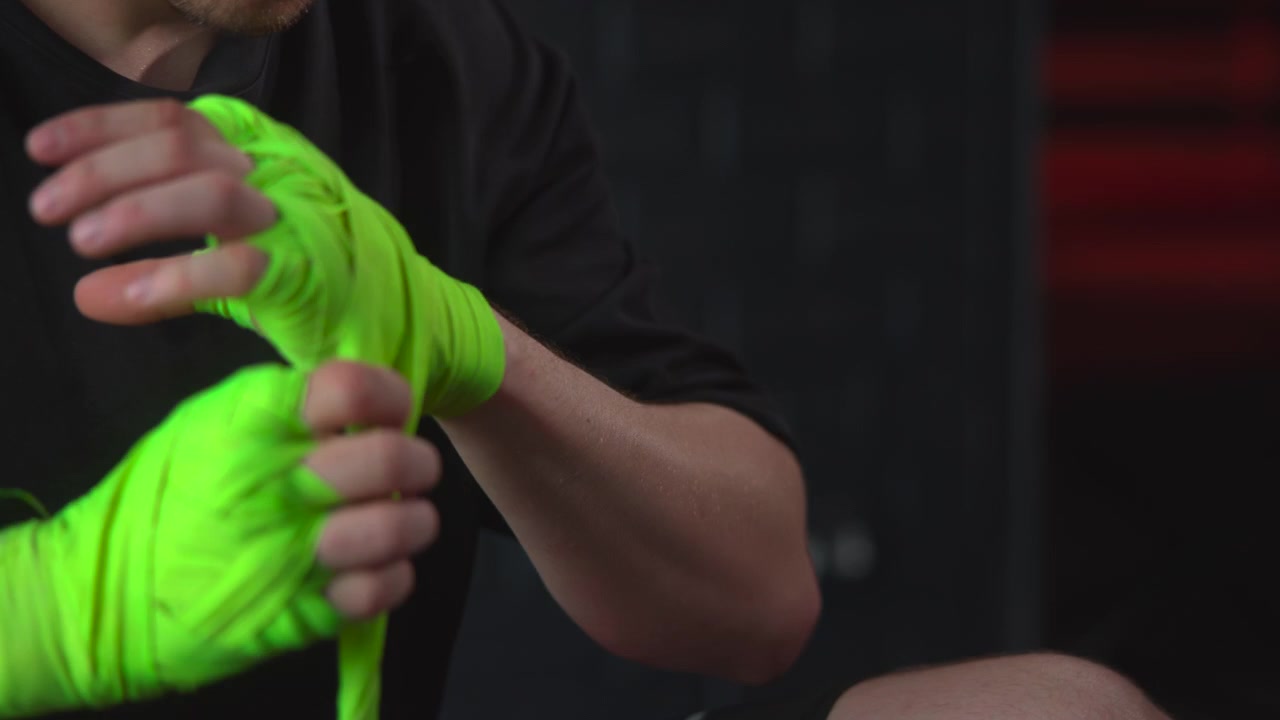 Primo piano mostra mani che avvolgono un supporto elastico verde neon brillante attorno a dita e polso, con pieghe a strati, tensione visibile, avambracci parziali e sfondo scuro sfocato per la preparazione all'allenamento.