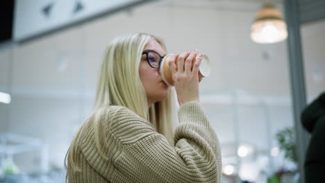 vista lateral de una joven con gafas bebiendo una bebida, mirando contemplativa en un centro comercial bien iluminado con iluminación elegante, ambiente suave y un transeúnte en el fondo