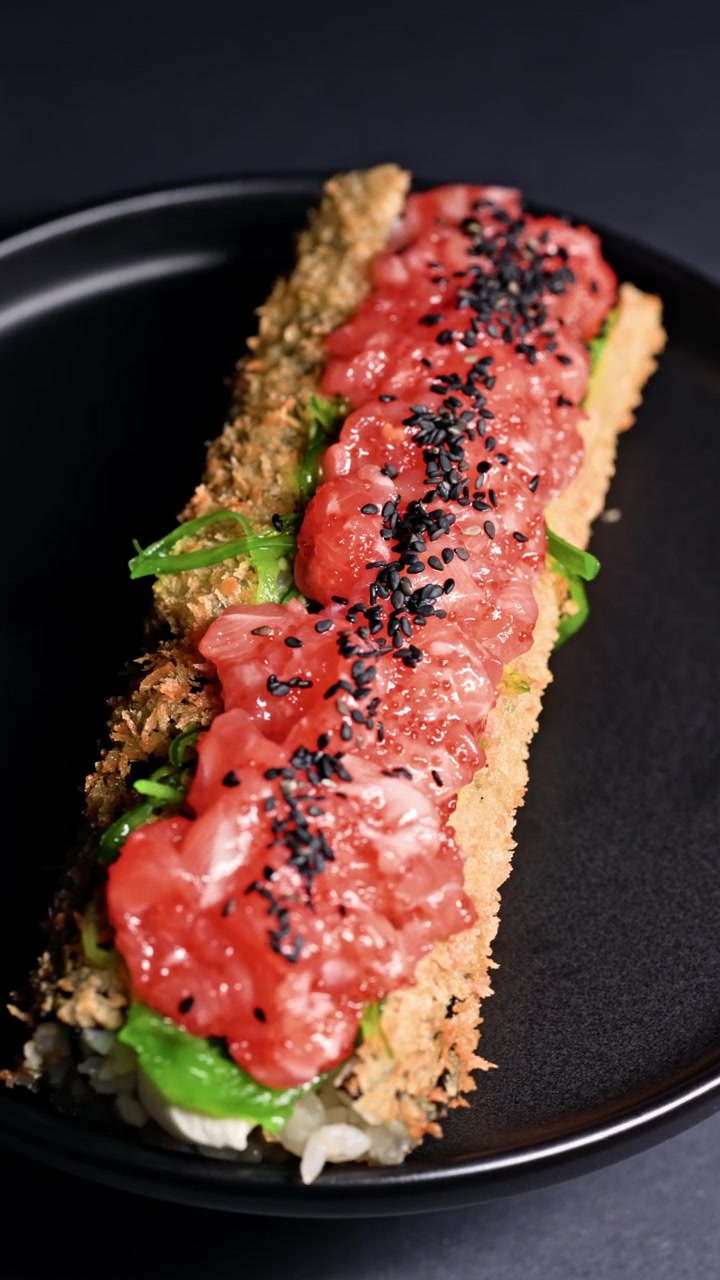 Rotolo di sushi con strato esterno fritto e guarnizione di pesce crudo su un piatto nero in un ristorante.
