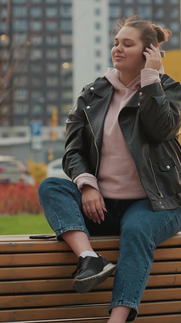 Female figure remains seated on bench wearing casual. Right hand holds headphones near ear as left arm rests. Surroundings include tall buildings parked cars trees grassy