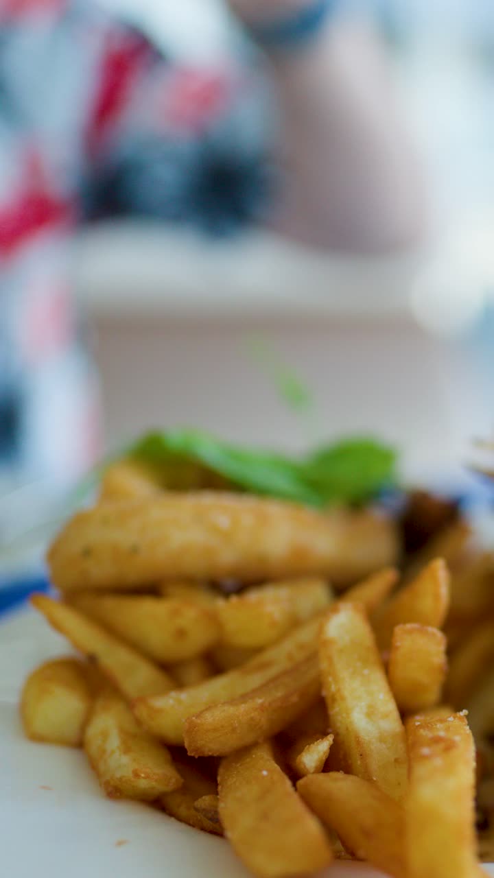 Golden fries and fried fish on plate, hand reaching, blurred background, natural daylight, shallow focus