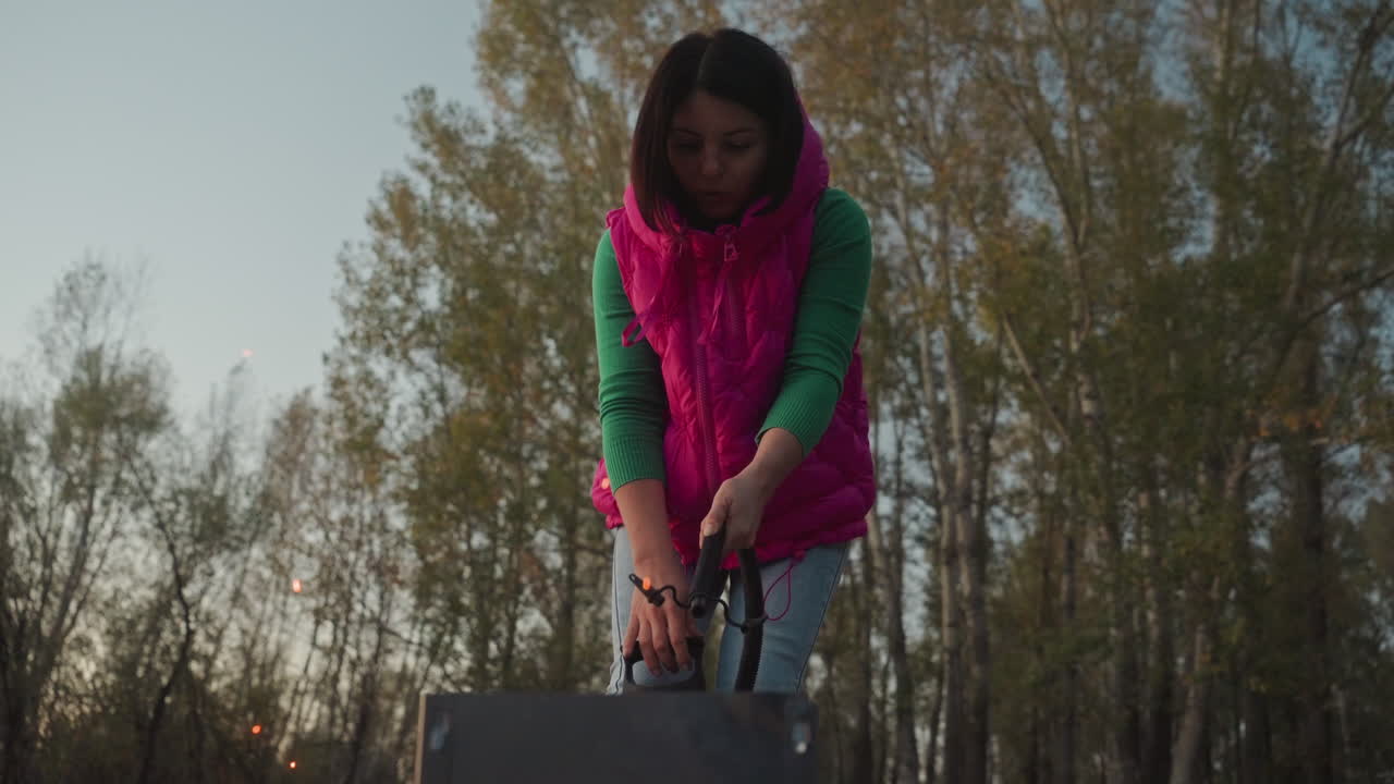 Donna in campeggio all'aperto che accende un fuoco su un fornello portatile al crepuscolo, con sciarpa rosa brillante e maglione verde, sfondo di foresta, scintille e bagliore di braci, scena di preparazione concentrata, atmosfera crepuscolare accogliente.