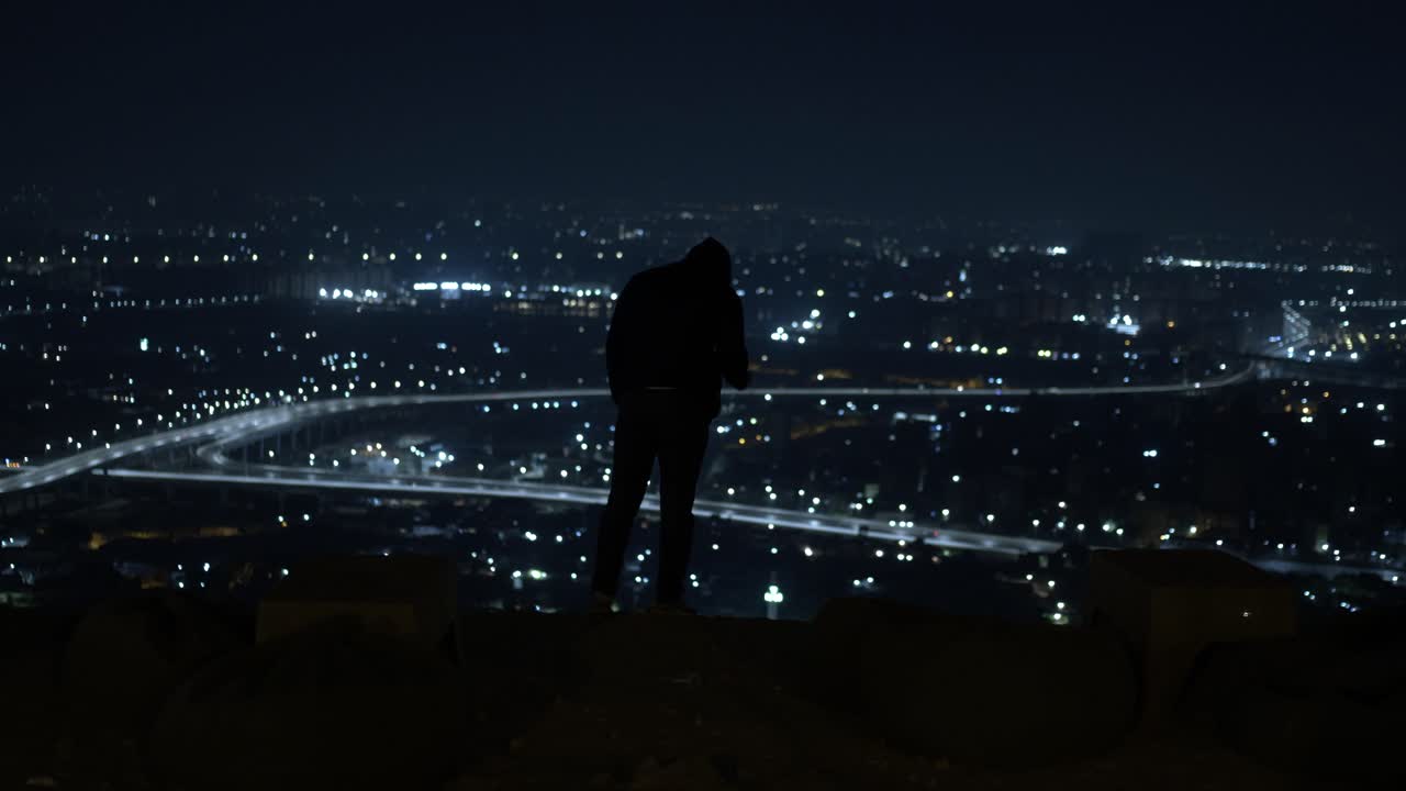 검은 재 ⁇ 을 입은 남자가 한밤중에 고속도로를 내려다보는 탑 뷰에 서 있습니다.