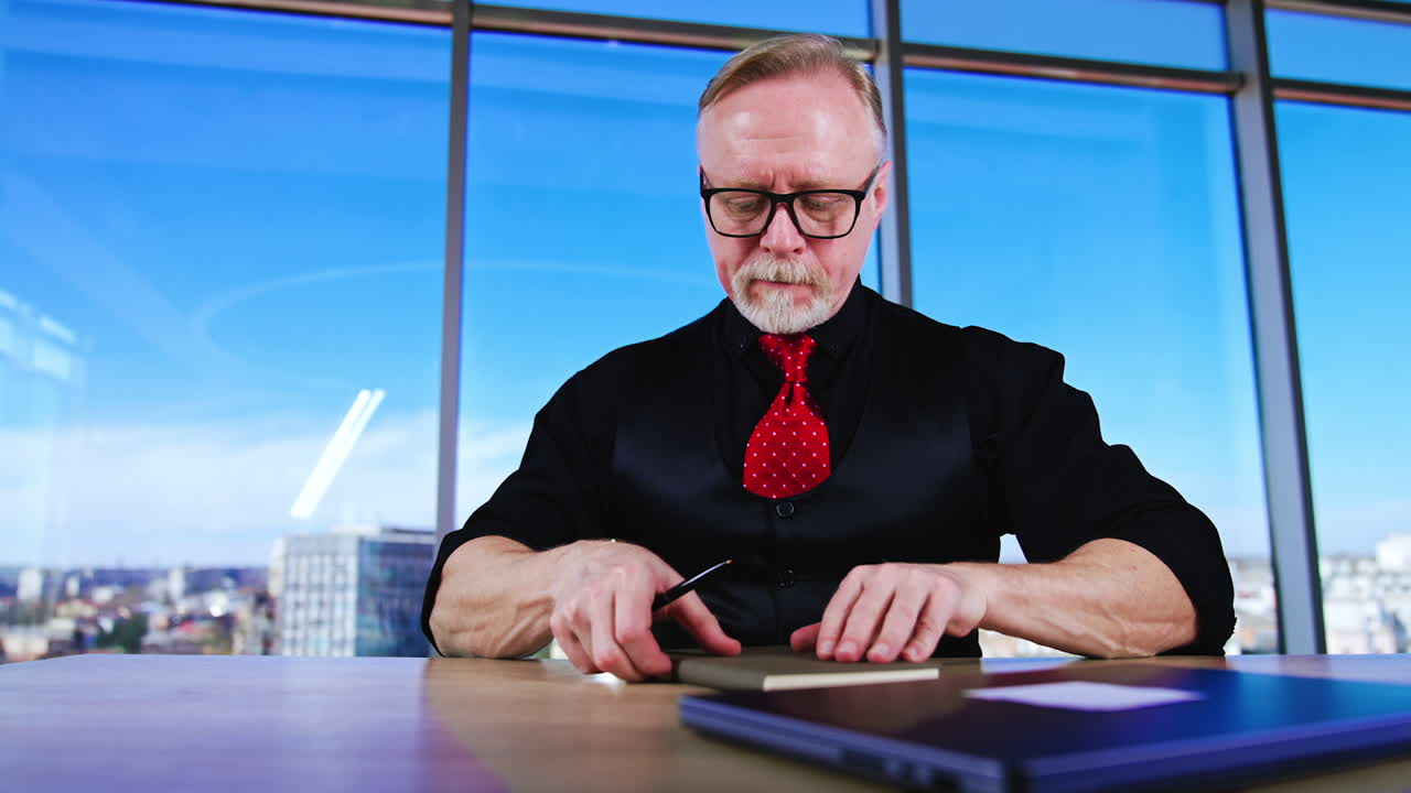 Smiling senior man in black shirt, vest and red tie sits at desk in office. Boss takes his paper book, opens laptop and starts work. Low angle view.
