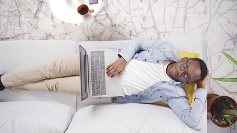african man lying on sofa at home resting and using laptop.