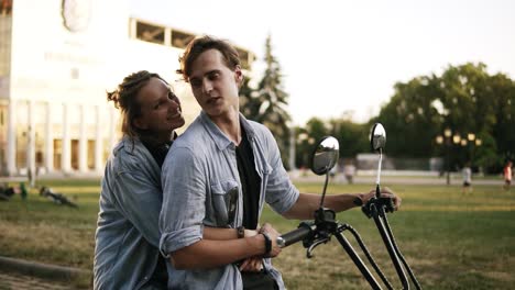 Overview-footage-of-a-beatuful-couple-sitting-on-a-bike.-Girl-is-hugging-man-from-the-back.-Wearing-black-T-shirt-and-blue-shirt.-Summer-day