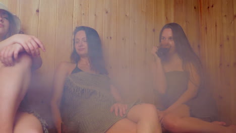 two women enjoy sauna session, one in foreground wears towel and felt hat with eyes closed in relaxation, second woman glances at her calmly while steam rises around them