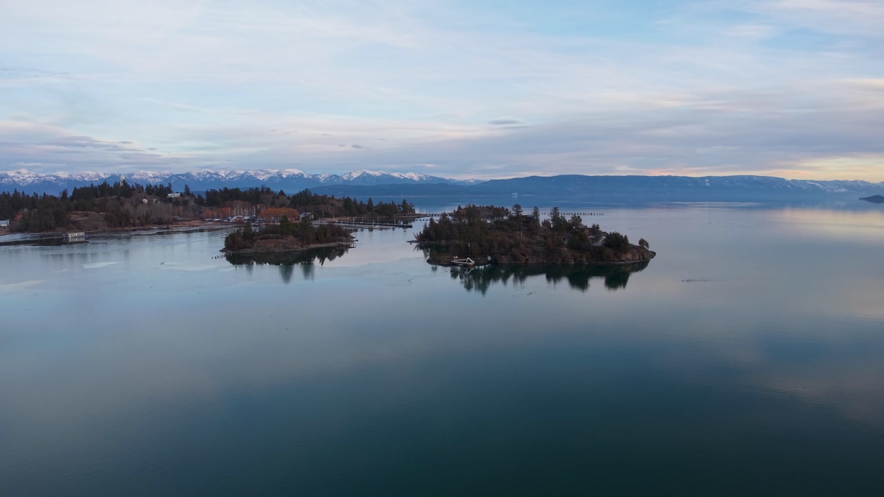 Aerial drone view of small tree-covered islands clustered together in a calm lake, with snowy mountains and a soft pastel sky in the background.