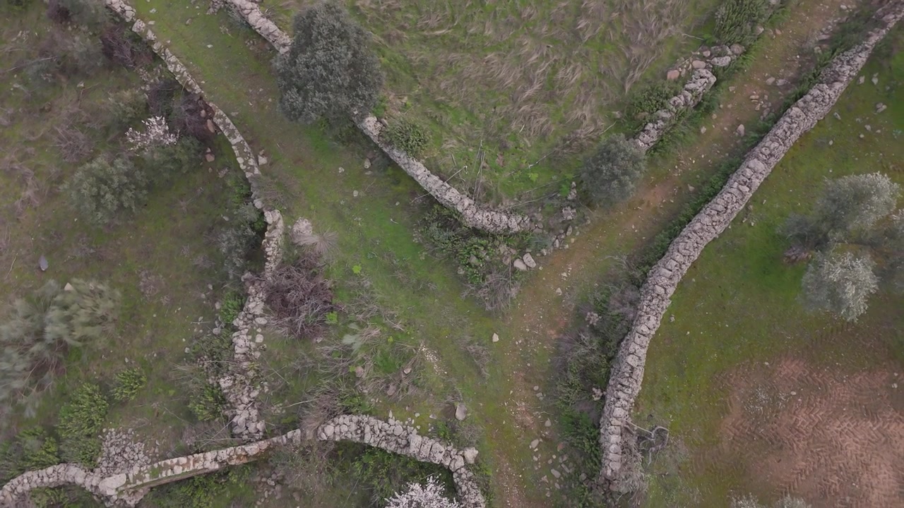 Slow motion top down drone ascent over a rural crossroads where curved stone walls from four adjoining properties meet, creating a striking geometric junction in the countryside.