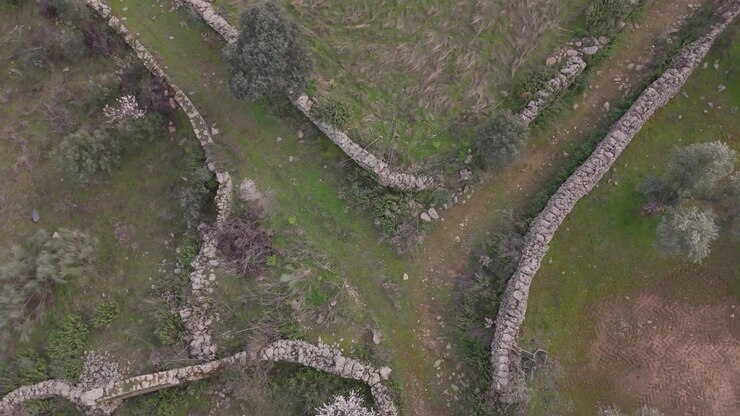 Slow motion top down drone ascent over a rural crossroads where curved stone walls from four adjoining properties meet, creating a striking geometric junction in the countryside.