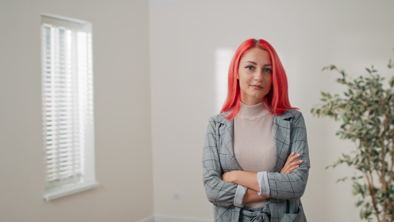 elegante mujer de cabello rosado está de pie contra la pared blanca en una habitación vacía, la chica tiene los brazos cruzados sobre el pecho ella sonríe a la cámara agente de bienes raíces asegurador ajustador de oficina corporativa trabajadora de negocios