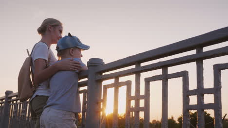 Una-Mujer-Con-Su-Hija-Está-De-Pie-En-La-Barandilla-Del-Puente-Al-Atardecer-Caminar-Con-Mamá