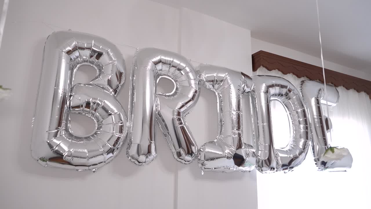 Ballonger i sølv former ordet «bride» i et lyst innendørs arbeidsområde. Dekorasjonene bidrar til den festlige stemningen før et bryllup.