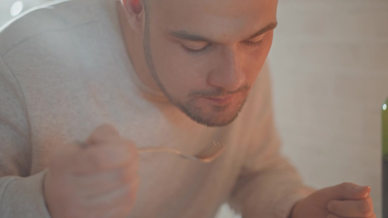 Chest up view of young man seated at table, twirling pasta with relaxed expression beside fruit plate and white bowl, warm indoor light, cozy home setting, casual mealtime atmosphere
