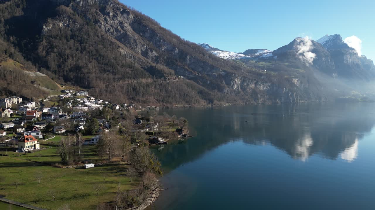 Clip con drone tranquilla che mostra un lago calmo, con un villaggio sulla riva e montagne alpine innevate sullo sfondo, in una soleggiata giornata primaverile