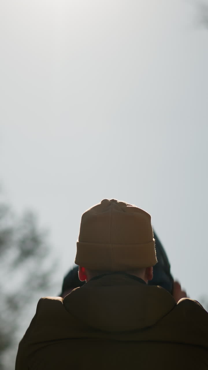 A father gently lowers his child from his shoulders, with a view of a bright sky and trees in the background