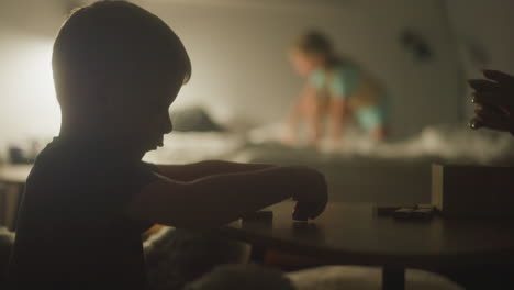 mom and little son build jenga tower from wooden blocks together with daughter playing around on bed on background. family spends evening playing board games