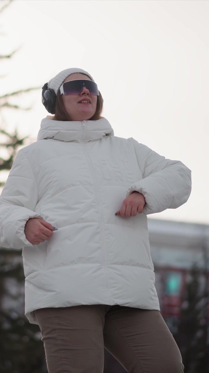 Donna con giacca invernale bianca, cuffie e occhiali da sole si muove energicamente all'aperto con freddo. Il suo linguaggio del corpo suggerisce che stia ballando o facendo esercizio a ritmo di musica mentre gesticola liberamente