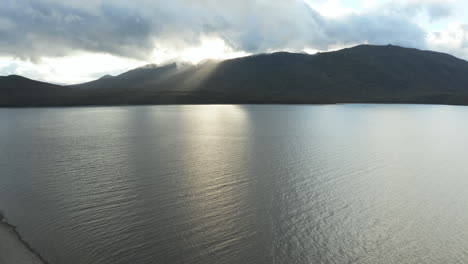 Fly-over-aerial-drone-footage-showcasing-rays-of-light-piercing-through-mountain-range-at-Lake-Te-Anau,-New-Zealand