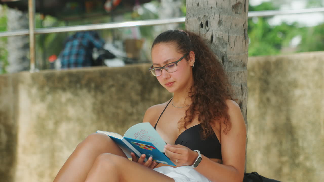 Woman reading book in swimsuit, sitting against tree in sunlit leisure, blue cover visible, relaxed vacation posture with sunglassesstyle glasses, flipping pages, tropical foliage and warm concrete