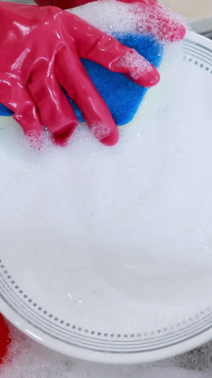 Hands in gloves wash a plate with a sponge in a kitchen sink filled with soapy water