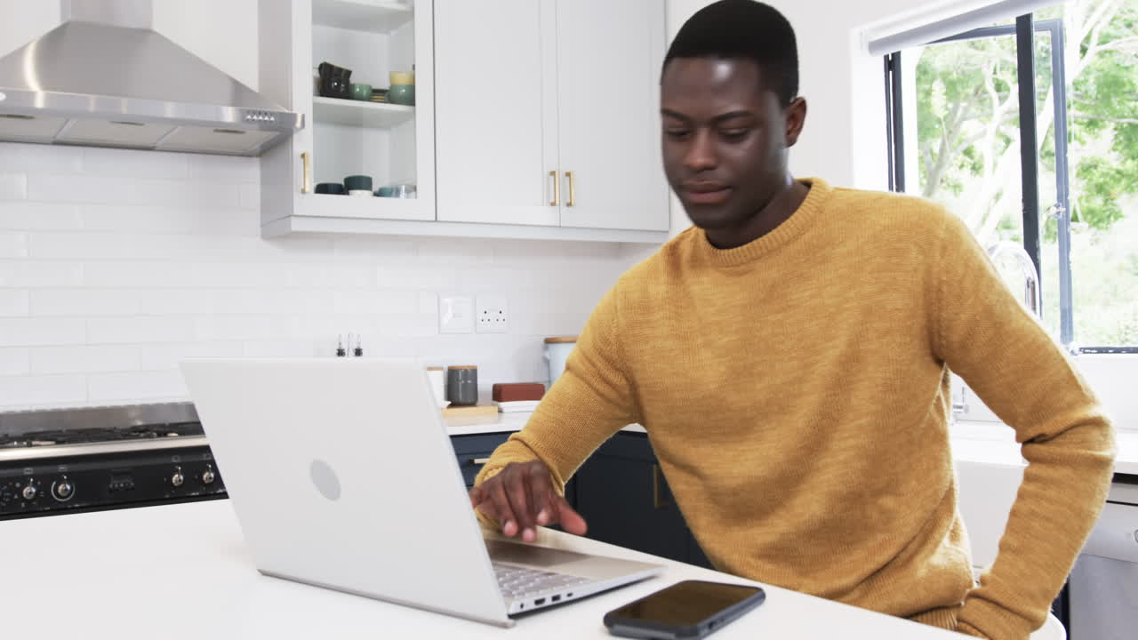 Man in kitchen using smartphone and laptop, wearing cozy sweater, working remotely, copy space