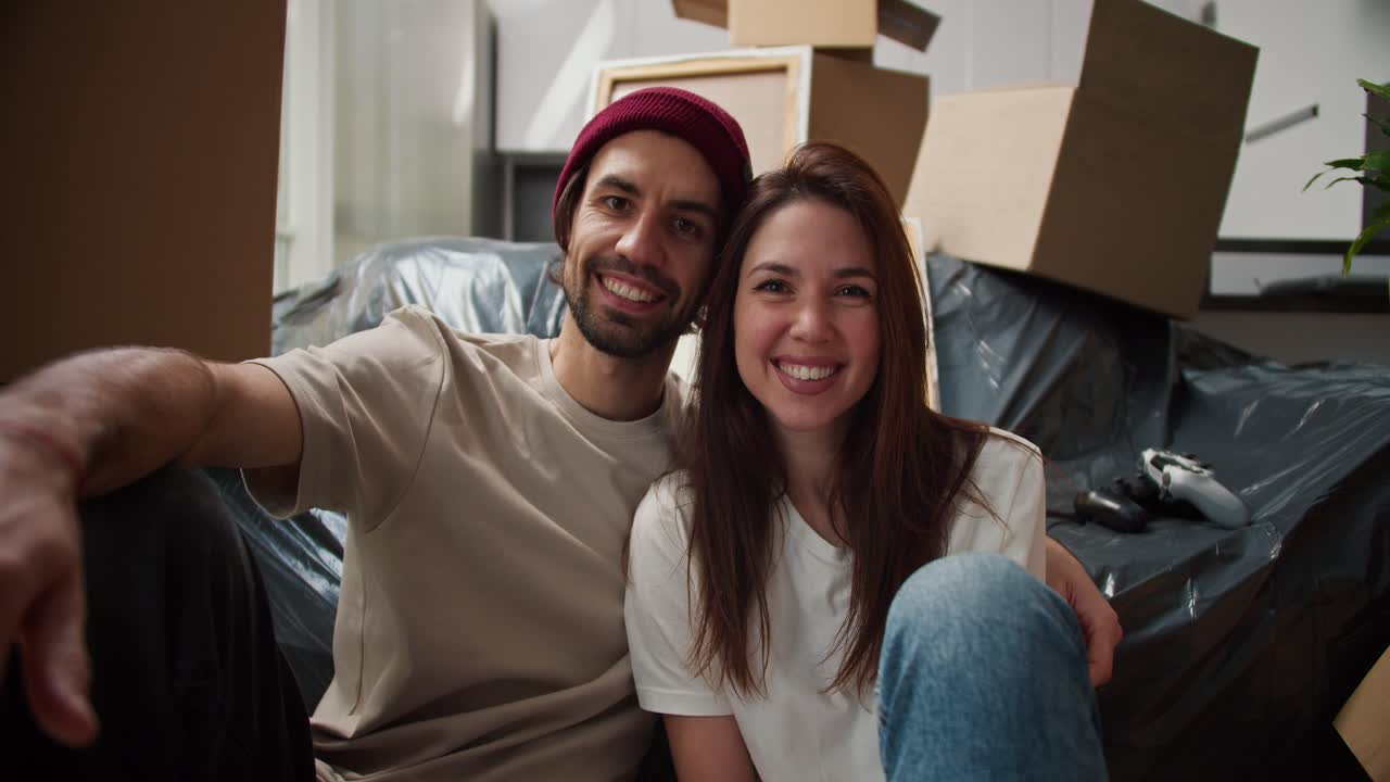Close-up portrait of a happy brunette man with stubble together with his brunette girlfriend in a white T-shirt they are sitting near the sofa packed in a black plastic case among a large number of boxes in a new apartment after moving