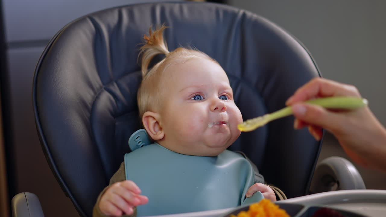 Adorable Caucasian baby sitting in a high chair wearing a bib. Mom feeds her child with a spoon.