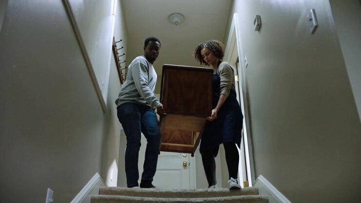 Lowering man in sweater and woman in apron stepping from landing, moving wood cabinet downstairs
