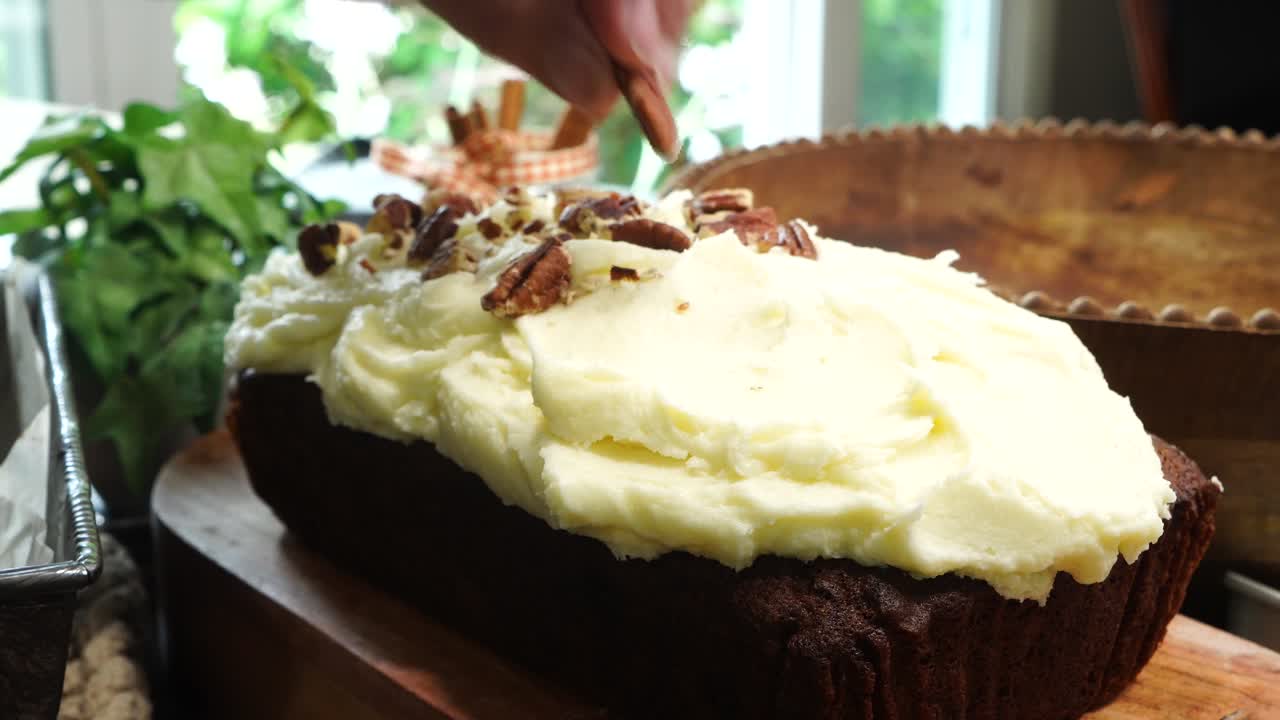 Baking a carrot cake loaf with cream cheese icing and topped with roasted salted pecans. Making a Autumn dessert with carrots, flour, sugar, nuts and frosting. Putting pecan or nuts on top of the cake