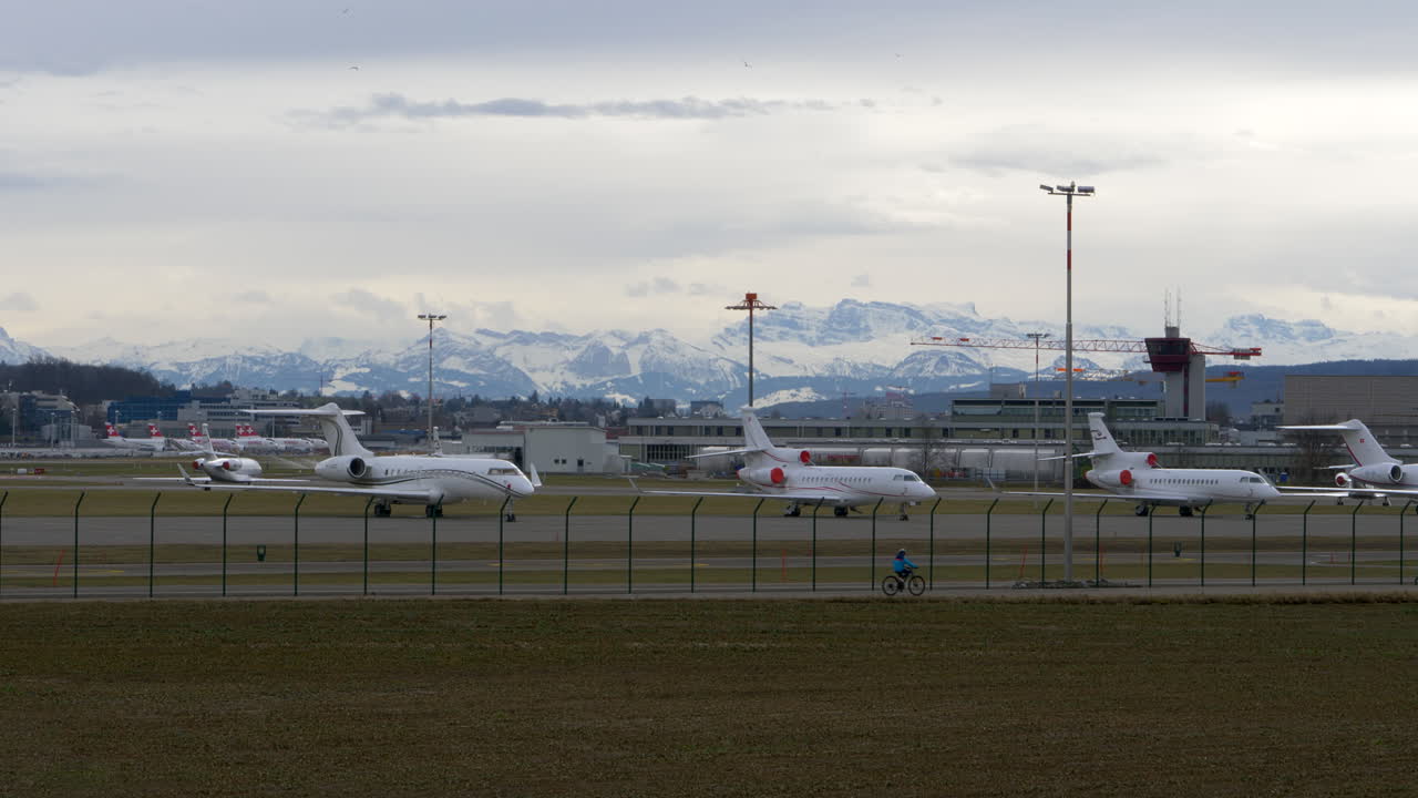 A line up of big corporate jets at an airport, white snowy mountains in the background.