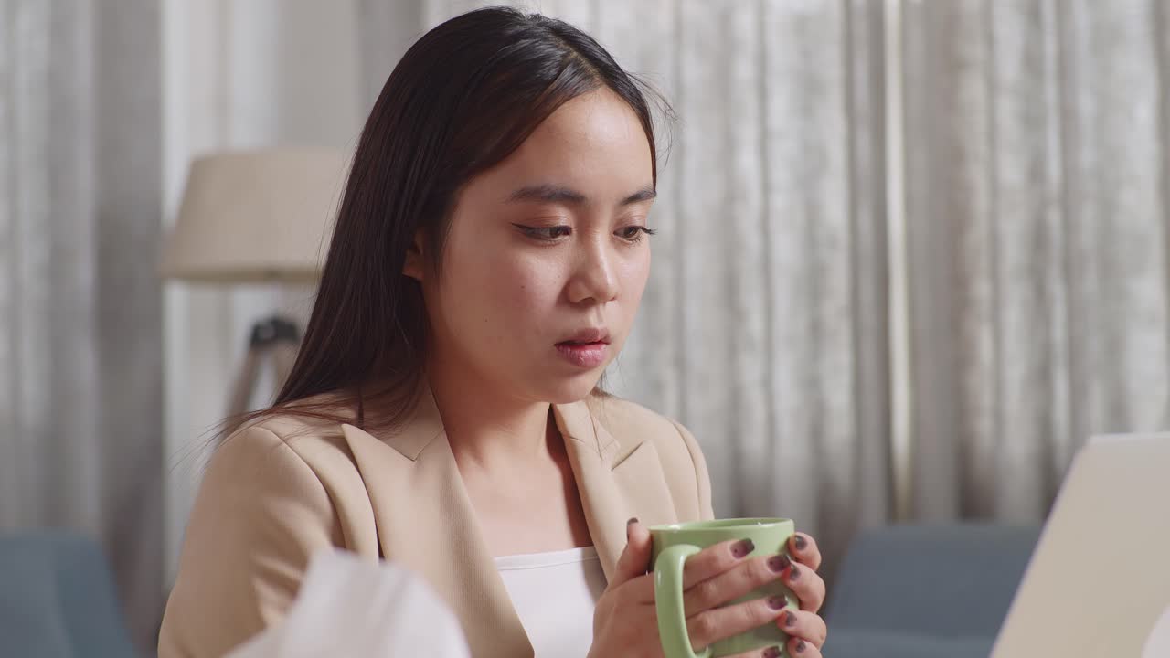 아픈 아시아 사업가 여성이 집에서 거실에서 노트북으로 일하면서 뜨거운 물을 마시고 있습니다.