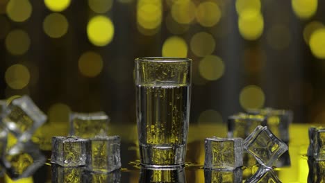 barman pour frozen vodka from bottle into shot glass. ice cubes against shiny gold party background