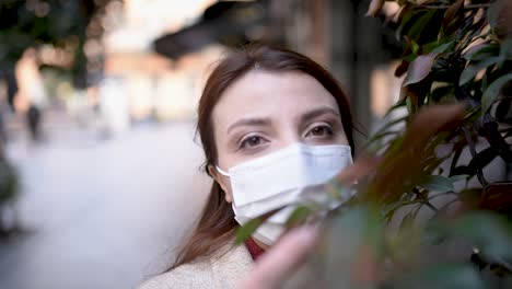 beautiful girl wearing protective medical mask and fashionable clothes looks at camera behind of plants