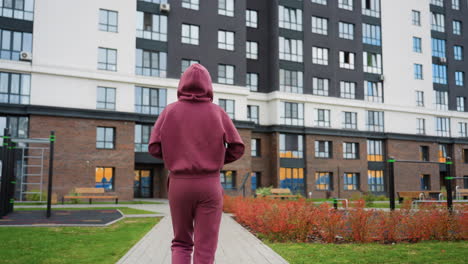 back view of fitness trainer in maroon hoodie walking along urban pavement toward tall highrise framed by autumn flowers, greenery and park bench capturing solo exercise routine in modern environment