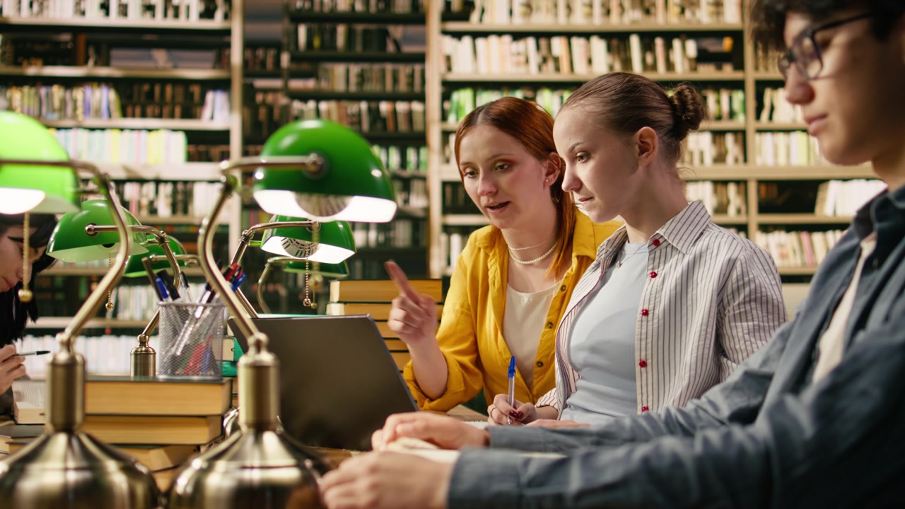 Tutor and student discuss research beside laptop and stacked books in library
