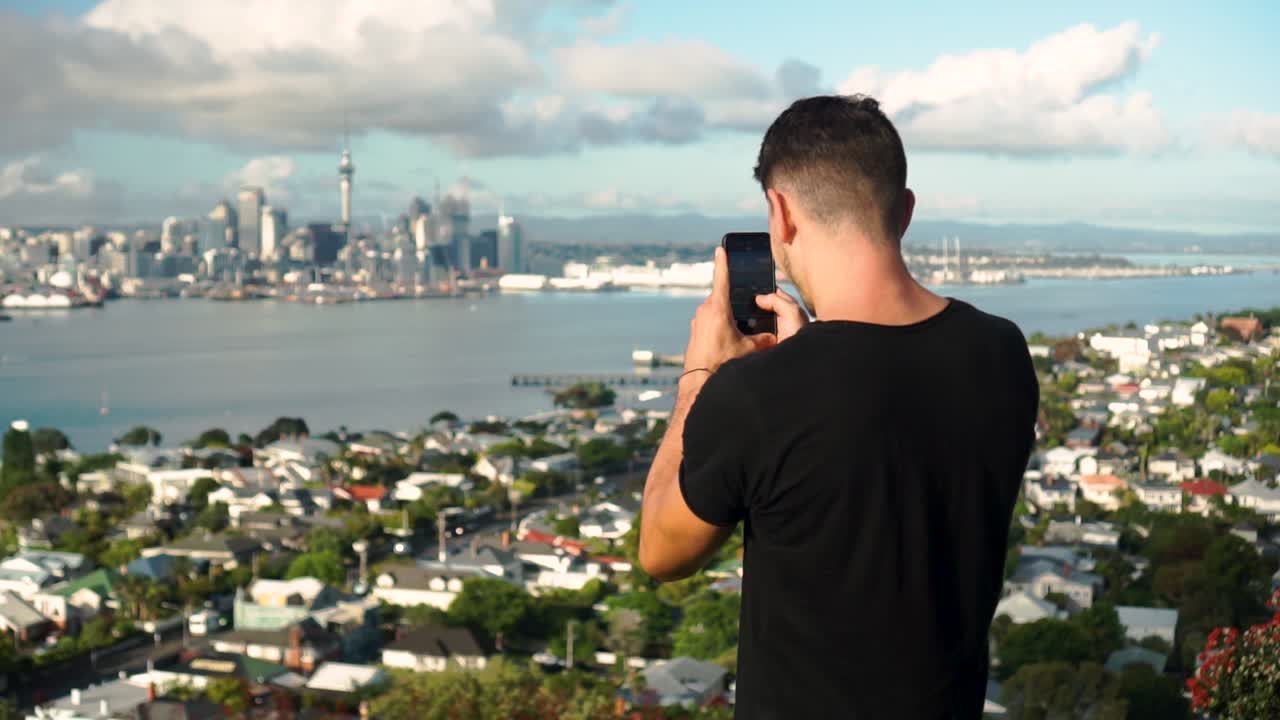 Young caucasian tourist taking a photo with phone of Auckland and Sky Tower from Mount Victoria, New Zealand