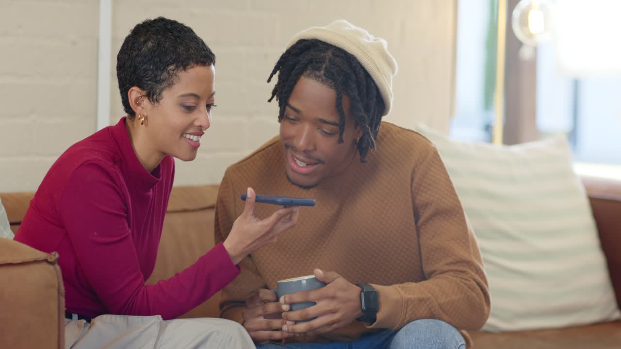 Couple holding coffee mug, speaking into smartphone while leaning closer to playback on beige sofa
