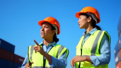 two construction workers in safety gear communicating at a shipping yard under clear blue skies, engaged in a task with containers in the background