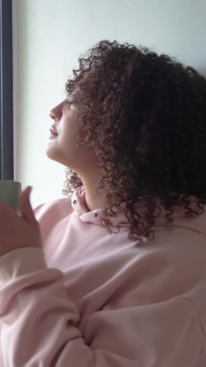 Video verticale: Donna in felpa rosa sorseggia da una tazza verde chiaro, appoggiata alla finestra con alberi, pensierosa.
