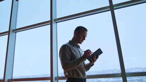 focused man standing in office with notebook in hands. businessman taking notes near the big window. cityscape at backdrop.